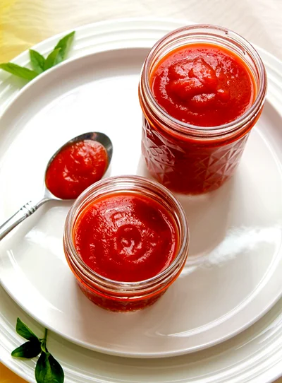 Two glass jars of thick homemade tomato ketchup and a spoonful of it on a white plate