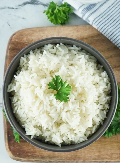 A grey bowl of fluffy steamed white rice topped with a sprig of parsley, on a wooden board