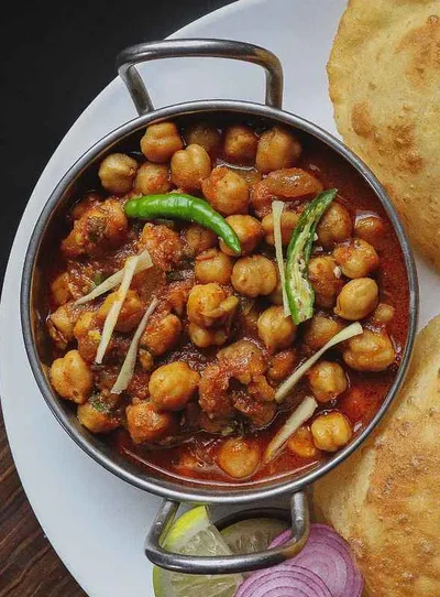 A steel kadhai of chole masala topped with green chilli and ginger strips, served with bhature, lime and onion